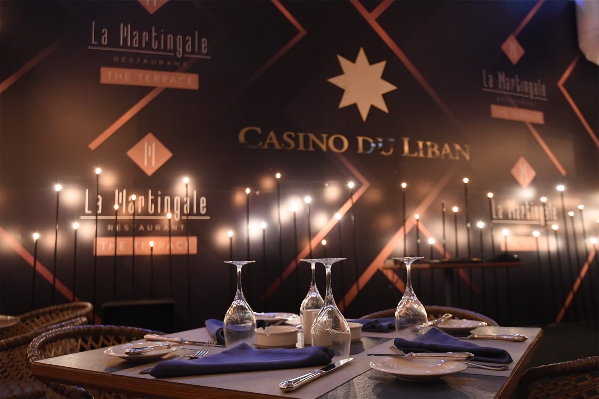A set dinner table on La Martingale terrace with upturned glasses, strings of small lights and the Casino du Liban star on the wall behind